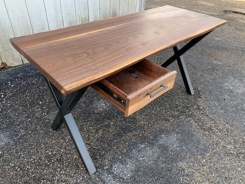 May include: A wooden desk with a live edge top and a single drawer. The desk has black metal legs in an X shape.