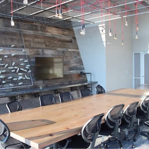 May include: A long wooden conference table with black mesh chairs in a modern office setting. The room has a rustic wood wall with a TV and exposed metal beams with hanging light bulbs.