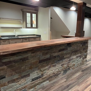 May include: A home bar featuring a live edge dark wood countertop and a base constructed from multi-colored stacked stone in shades of gray, tan, and brown. Behind the bar, a kitchen area includes light wood cabinets, floating shelves, and a sink.