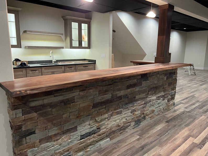 May include: A home bar featuring a live edge dark wood countertop and a base constructed from multi-colored stacked stone in shades of gray, tan, and brown. Behind the bar, a kitchen area includes light wood cabinets, floating shelves, and a sink.