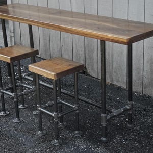 May include: A wooden bar table and two stools. The table has a rectangular top and is supported by black metal pipes. The stools have square wooden seats and are also supported by black metal pipes. The wood has a warm, natural finish.