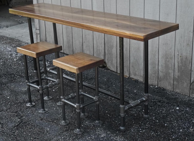 May include: A wooden bar table and two stools. The table has a rectangular top and is supported by black metal pipes. The stools have square wooden seats and are also supported by black metal pipes. The wood has a warm, natural finish.