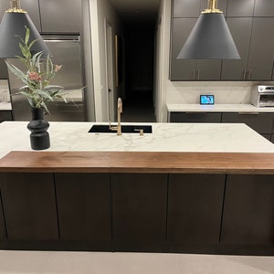 May include: A modern kitchen with dark gray cabinets, a white countertop, and a large island. A wooden extension is attached to the island. A black vase with flowers sits on the countertop. Two black pendant lights hang above the island.