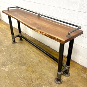May include: A rustic console table with a live edge wood top. The table has black metal pipe legs and a matching railing along the top. The table is approximately 152 cm long, 38 cm wide, and 76 cm high.