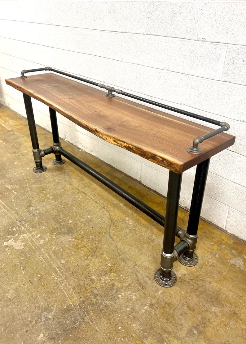 May include: A rustic console table with a live edge wood top. The table has black metal pipe legs and a matching railing along the top. The table is approximately 152 cm long, 38 cm wide, and 76 cm high.