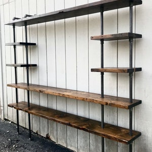 May include: A wall-mounted industrial-style shelf unit with multiple wooden shelves supported by black metal pipes. The shelves are made of dark stained wood and the unit is mounted on a white wall.