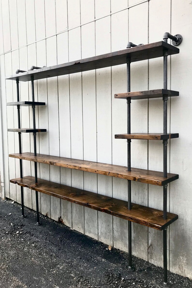 May include: A wall-mounted industrial-style shelf unit with multiple wooden shelves supported by black metal pipes. The shelves are made of dark stained wood and the unit is mounted on a white wall.