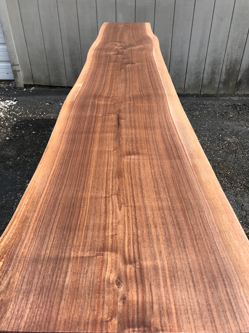May include: A long, rectangular slab of natural walnut wood featuring a prominent live edge along the side. The surface displays a rich brown color with visible wood grain patterns, knots, and variations in tone throughout the length of the board.