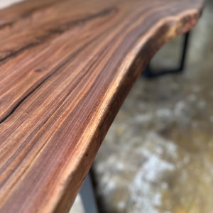May include: A live edge table featuring a dark brown walnut wood slab with a prominent natural grain pattern and smooth finish, supported by black metal legs. The surface displays deep brown and reddish tones with visible wood texture.
