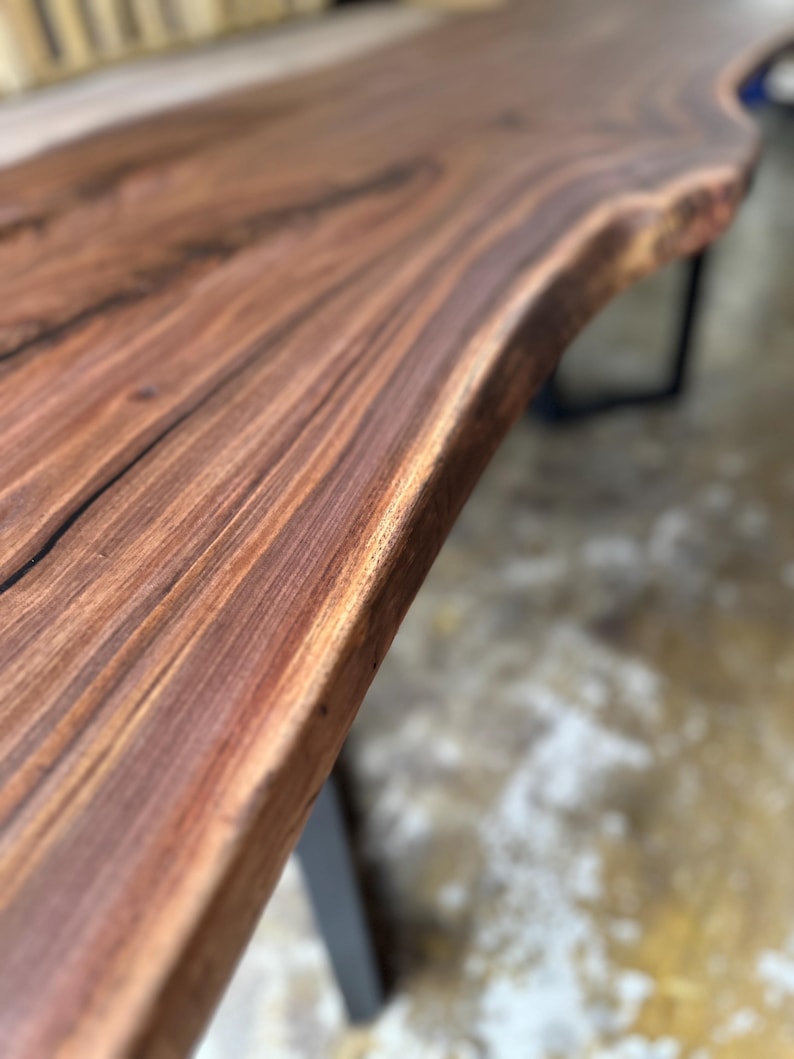 May include: A live edge table featuring a dark brown walnut wood slab with a prominent natural grain pattern and smooth finish, supported by black metal legs. The surface displays deep brown and reddish tones with visible wood texture.