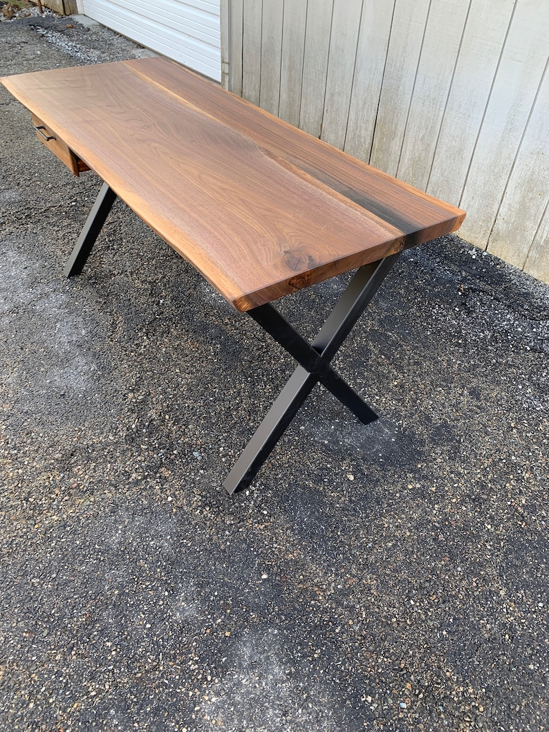 May include: A dark brown wooden desk with a live edge top and a single drawer. The desk has black metal X-shaped legs.