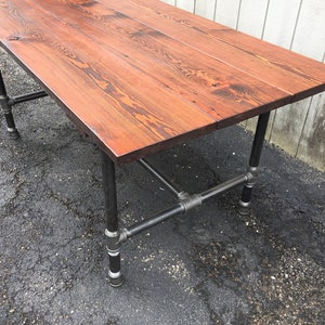 May include: A rectangular wooden table with a reddish-brown finish. The tabletop is supported by black metal pipe legs and crossbars, creating an industrial aesthetic. The table's dimensions are not visible.