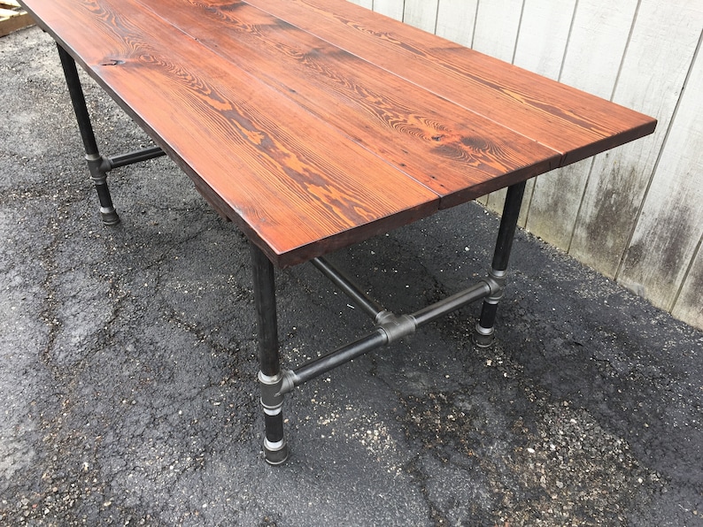 May include: A rectangular wooden table with a reddish-brown finish. The tabletop is supported by black metal pipe legs and crossbars, creating an industrial aesthetic. The table's dimensions are not visible.