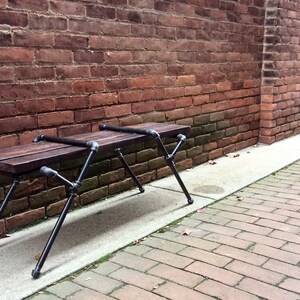 May include: A wooden bench with black metal legs sits in front of a brick wall. The bench is made of reclaimed wood and has a rustic look.