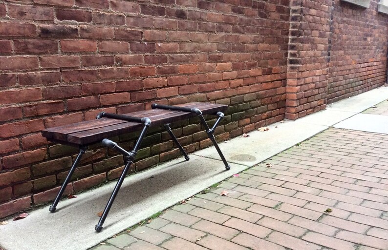 May include: A wooden bench with black metal legs sits in front of a brick wall. The bench is made of reclaimed wood and has a rustic look.
