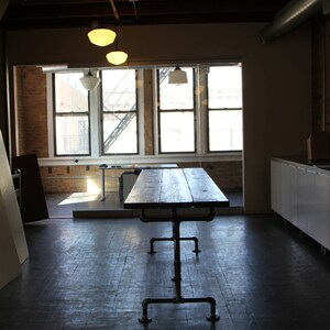 May include: A dark wood table with a metal pipe frame stands in an open space with large windows. The table is the focal point, with a rustic, industrial design. Overhead, three pendant lights illuminate the room. The floor is dark wood.