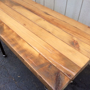 May include: A rectangular wooden table with a natural finish. The tabletop is made from multiple wooden planks, showing the grain and knots. The table is supported by black metal legs. The wood has a warm, light brown colour.