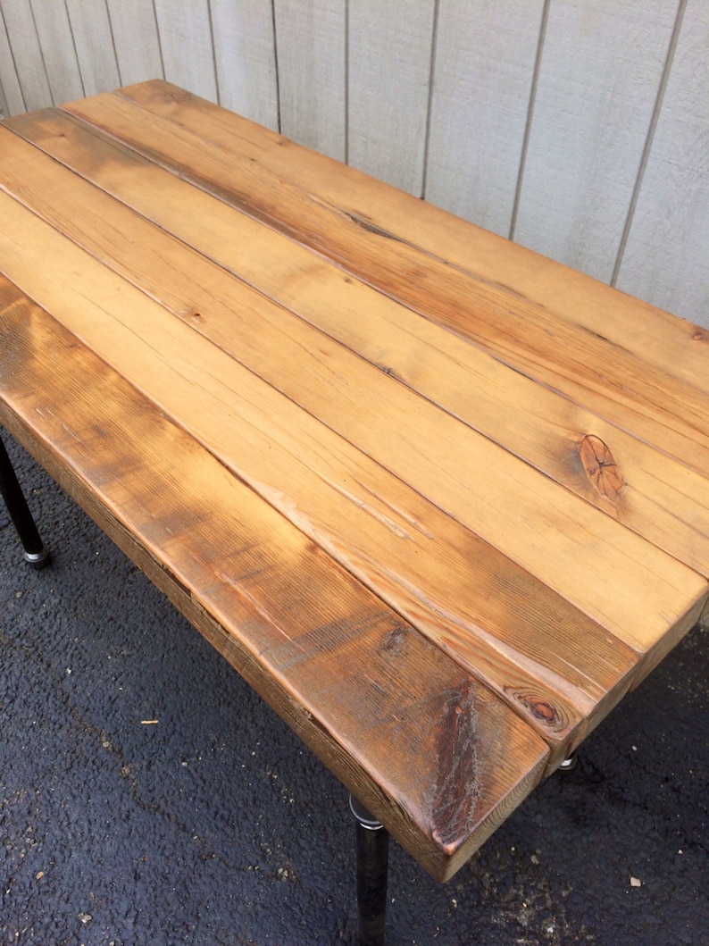 May include: A rectangular wooden table with a natural finish. The tabletop is made from multiple wooden planks, showing the grain and knots. The table is supported by black metal legs. The wood has a warm, light brown colour.