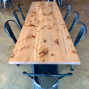 May include: A long, wooden table with a natural finish sits on a concrete floor.  Eight black metal chairs surround the table.