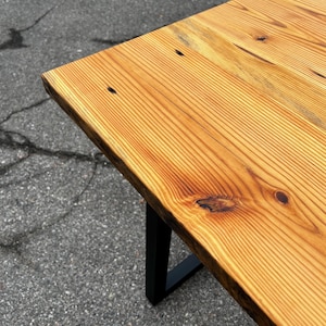 May include: A wooden table with a natural wood grain finish and a black metal leg. The table's top is made of light brown wood with visible knots and grain patterns. The table is approximately 76 cm long.