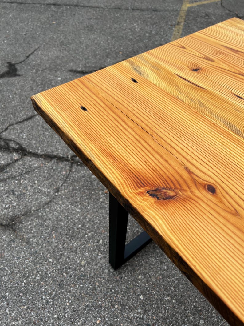 May include: A wooden table with a natural wood grain finish and a black metal leg. The table's top is made of light brown wood with visible knots and grain patterns. The table is approximately 76 cm long.