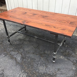 May include: A rectangular wooden desk with a reddish-brown finish. The tabletop is supported by black metal pipes and fittings, creating an industrial aesthetic. The desk is approximately 152 cm long.