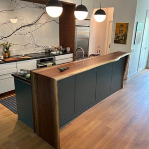 May include: A kitchen island featuring a dark wood waterfall countertop with a natural live edge, paired with dark gray cabinetry. Three spherical pendant lights with black tops and white glass globes hang above the island in a modern kitchen.