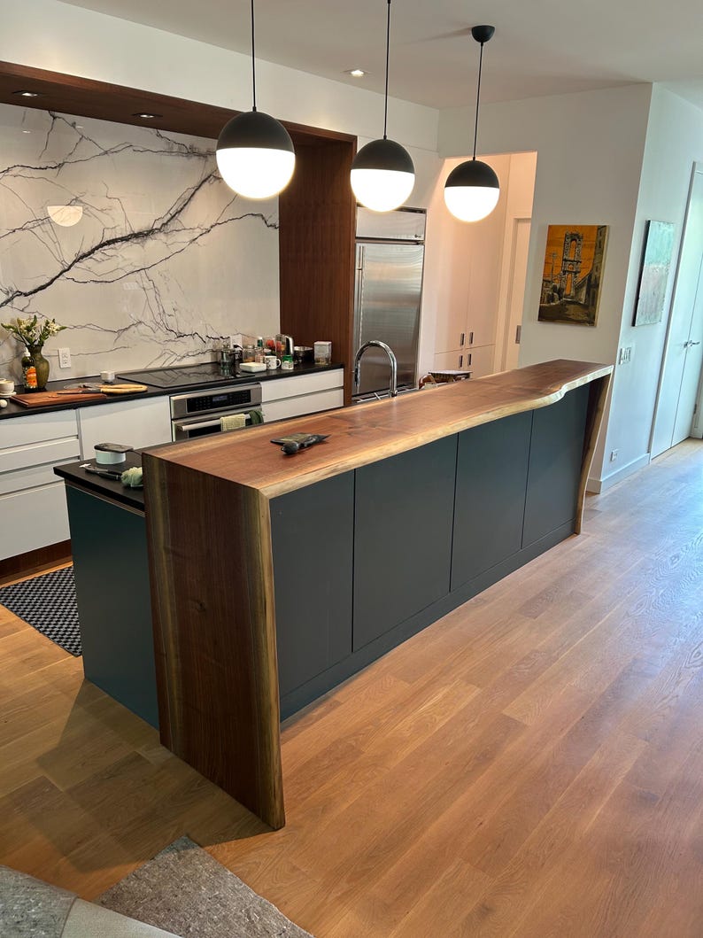 May include: A kitchen island featuring a dark wood waterfall countertop with a natural live edge, paired with dark gray cabinetry. Three spherical pendant lights with black tops and white glass globes hang above the island in a modern kitchen.