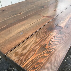 May include: A close-up of a wooden table with a dark brown stain. The table is made from several planks of wood, showing the natural grain and knots. The table's edges are square, and the legs are made of metal pipes.