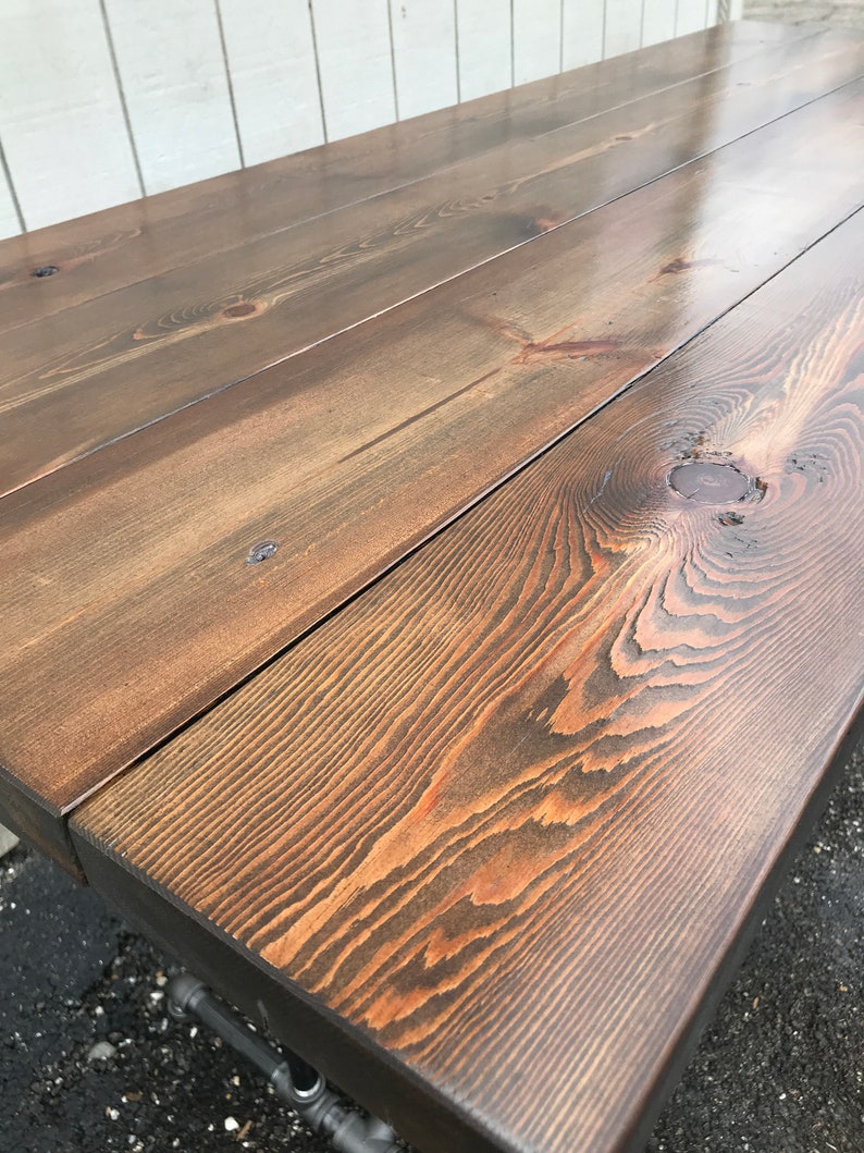 May include: A close-up of a wooden table with a dark brown stain. The table is made from several planks of wood, showing the natural grain and knots. The table's edges are square, and the legs are made of metal pipes.