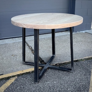 May include: A round wooden table with a whitewashed finish and a black metal base. The table has a simple, modern design and would be a great addition to any home.