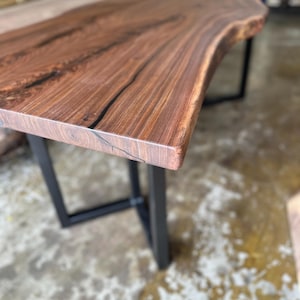 May include: A rectangular table featuring a dark brown wood slab top with a natural live edge and visible grain patterns. The table is supported by two black metal U-shaped legs positioned at each end of the wooden surface.