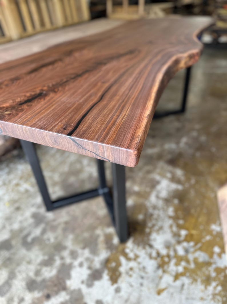 May include: A rectangular table featuring a dark brown wood slab top with a natural live edge and visible grain patterns. The table is supported by two black metal U-shaped legs positioned at each end of the wooden surface.