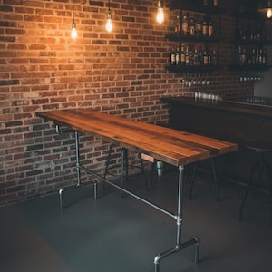 May include: A long, rectangular wooden table with a natural finish, supported by a metal pipe frame. The table is in a dimly lit room with a brick wall background and hanging light bulbs. The table is approximately 8 feet long.