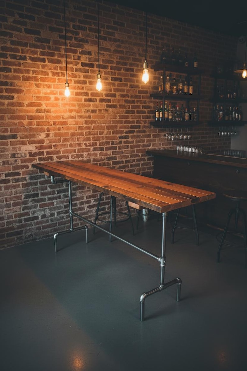 May include: A long, rectangular wooden table with a natural finish, supported by a metal pipe frame. The table is in a dimly lit room with a brick wall background and hanging light bulbs. The table is approximately 8 feet long.