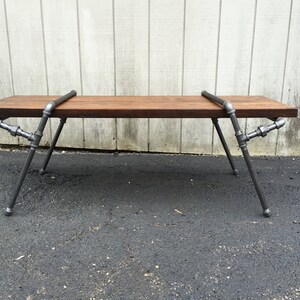 May include: A rustic wooden bench with a dark brown stain, supported by a metal frame made of industrial pipes. The bench has a simple design and a sturdy construction.