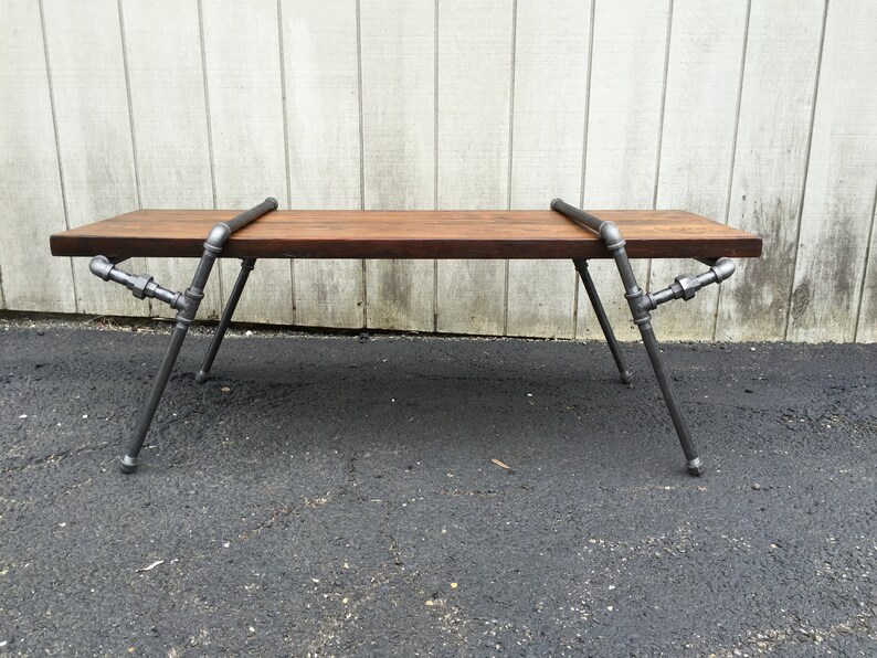 May include: A rustic wooden bench with a dark brown stain, supported by a metal frame made of industrial pipes. The bench has a simple design and a sturdy construction.