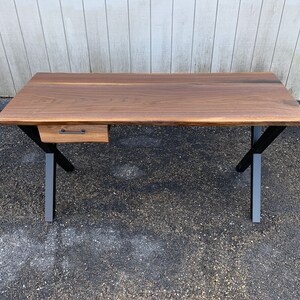 May include: A walnut wood desk with a single drawer and black metal legs. The desk has a live edge design and a natural wood finish.