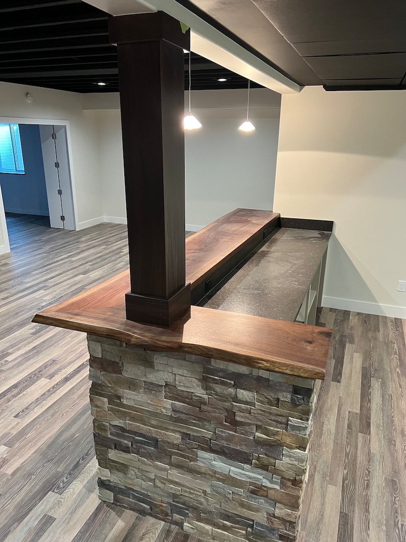 May include: A kitchen island featuring a live-edge dark wood countertop and a stone veneer base. A square dark wood support column rises from the center of the counter. The workspace includes a dark gray textured surface and integrated electrical outlets.
