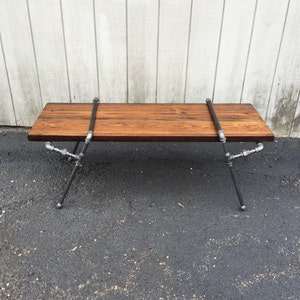 May include: A long, wooden bench with a dark brown stain. The bench is supported by black metal pipes that form an X shape on each end. The bench is sitting on a grey asphalt surface.