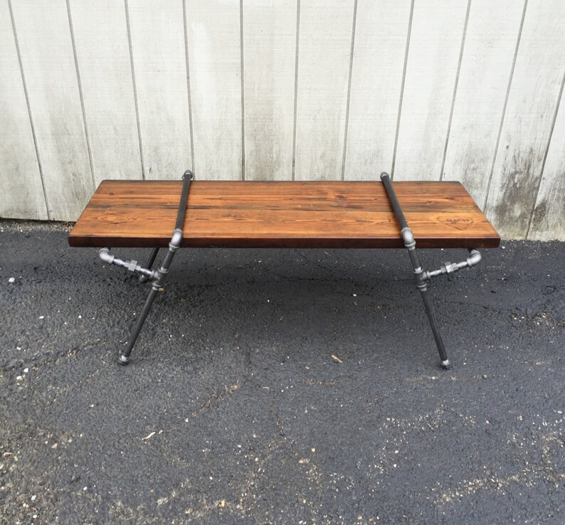 May include: A long, wooden bench with a dark brown stain. The bench is supported by black metal pipes that form an X shape on each end. The bench is sitting on a grey asphalt surface.