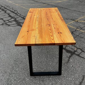 May include: A rectangular wooden table with a natural wood finish and black metal legs. The tabletop is made of planks of wood, showcasing the natural grain and knots. The table is approximately 183 cm long.