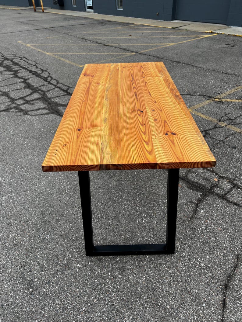 May include: A rectangular wooden table with a natural wood finish and black metal legs. The tabletop is made of planks of wood, showcasing the natural grain and knots. The table is approximately 183 cm long.