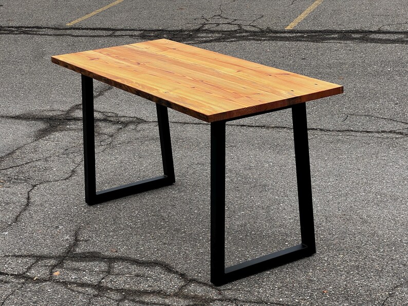 May include: A rectangular wooden table with a light brown surface and black metal legs. The table is approximately 152 cm long and 76 cm wide, with a simple, modern design. The legs are a square shape.