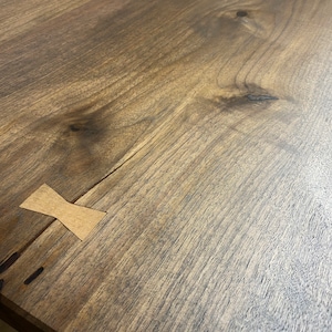 May include: A dark brown wooden table surface featuring a prominent natural wood grain pattern with visible knots and a light-colored, bow-tie shaped wooden inlay spanning across a thin crack in the wood to provide structural reinforcement.