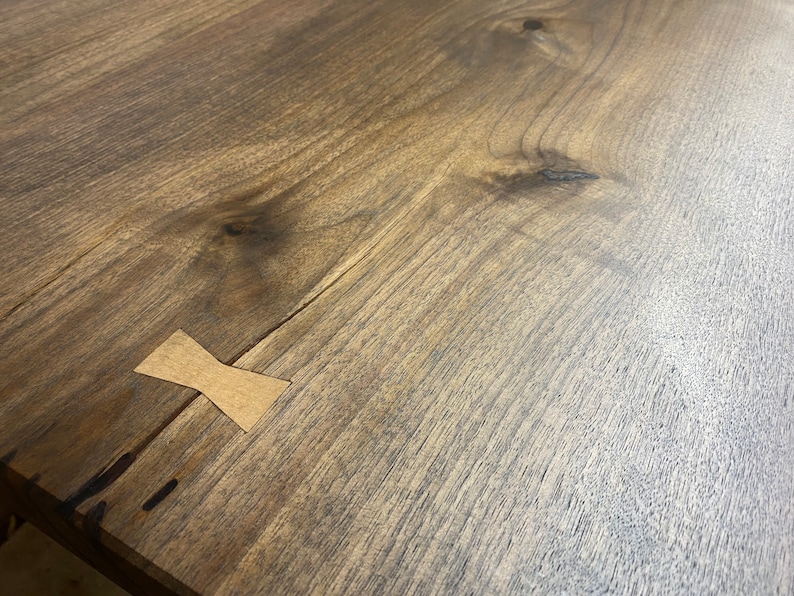 May include: A dark brown wooden table surface featuring a prominent natural wood grain pattern with visible knots and a light-colored, bow-tie shaped wooden inlay spanning across a thin crack in the wood to provide structural reinforcement.