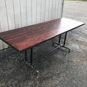 May include: A rectangular wooden table with a dark reddish-brown finish. The table is supported by black metal pipe legs. The table is approximately 72 inches long and 36 inches wide.