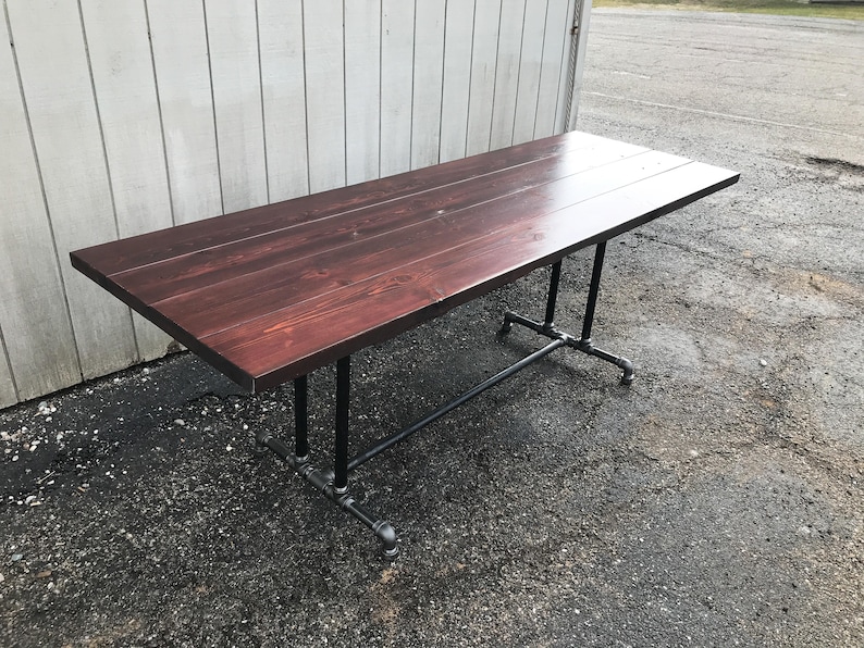 May include: A rectangular wooden table with a dark reddish-brown finish. The table is supported by black metal pipe legs. The table is approximately 72 inches long and 36 inches wide.