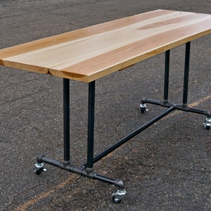 May include: A rectangular wooden table with a light brown surface and black metal legs. The table has a natural wood grain pattern and is mounted on wheels for mobility. The table is suitable for various uses.