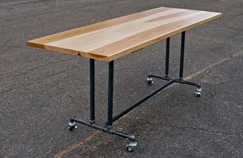 May include: A rectangular wooden table with a light brown surface and black metal legs. The table has a natural wood grain pattern and is mounted on wheels for mobility. The table is suitable for various uses.
